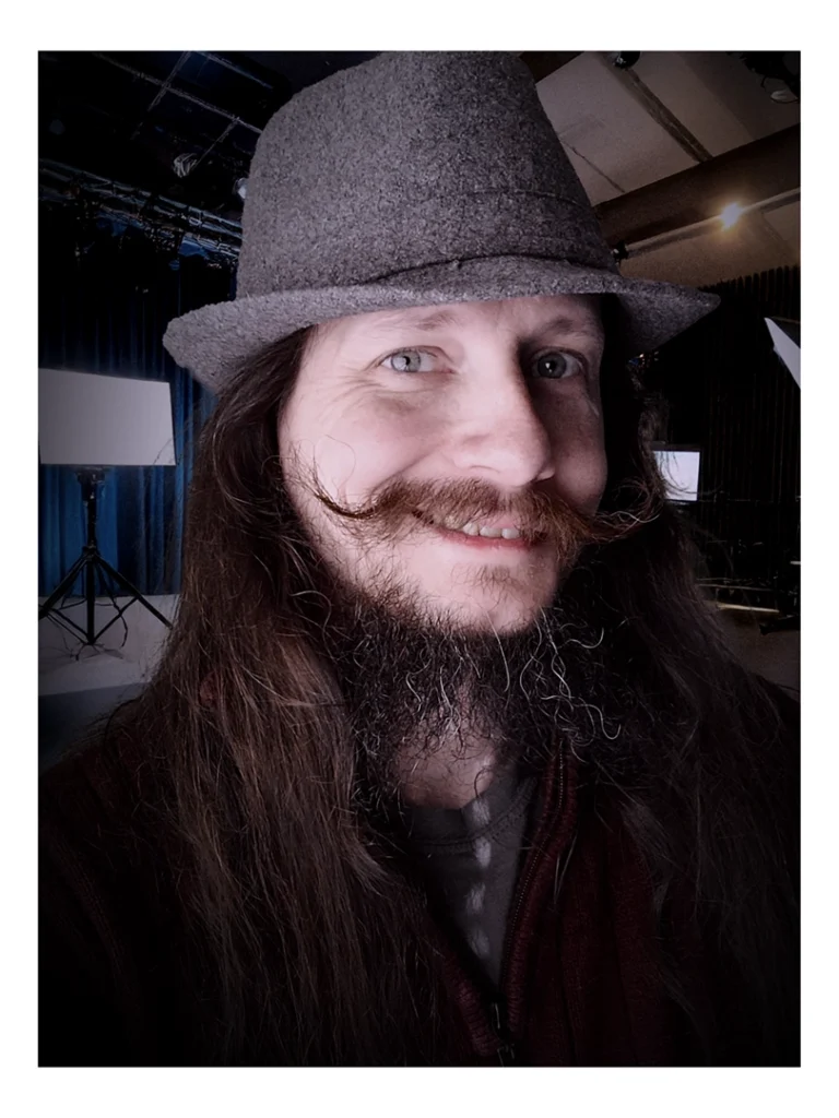 Owndog in a studio looking at the camera and smiling. He is wearing a red sweater and gray trilby.